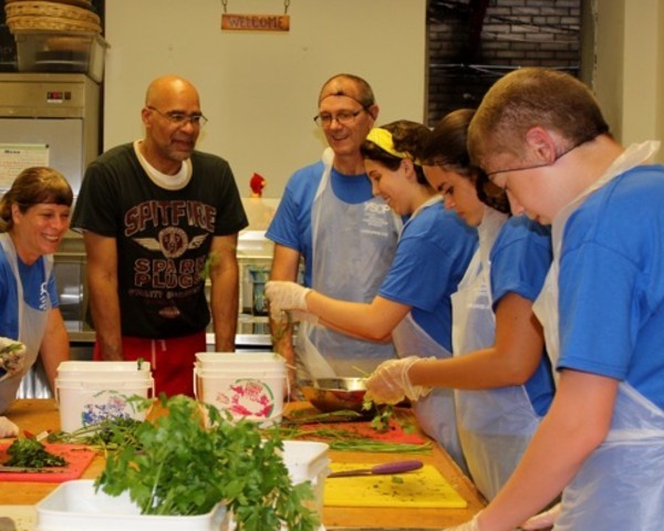 Volunteers preparing food for a meal service (Photo credit: Broadway Community) Six people around a large counter preparing food on cutting boards.