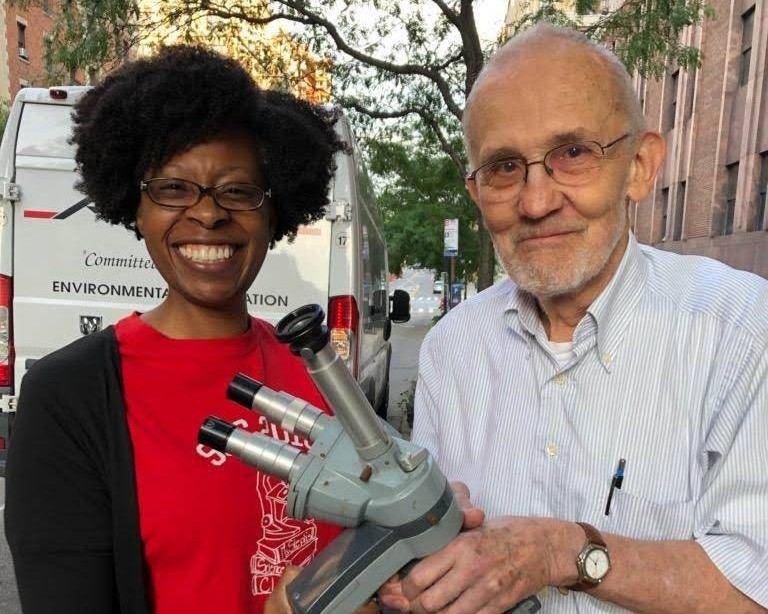African American Woman in Red and Black Shirt poses with Elderly White Man - both holding a research microscope.
